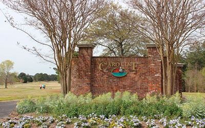 A red brick community entrance sign for Caroline with a decorative emblem, surrounded by trees and flowers.