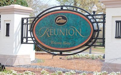 A decorative, oval Reunion: Welcome Home sign mounted between two white pillars in a landscaped setting.