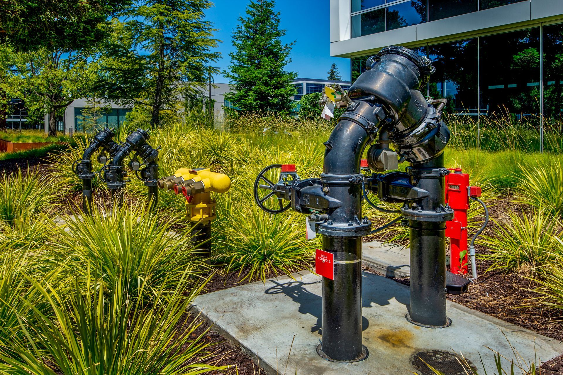 Industrial black water pipes with valves and a red fire hydrant stand on a concrete pad surrounded by green landscaping.