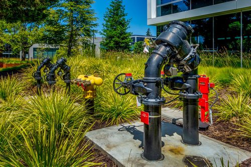 Industrial black water pipes with valves and a red fire hydrant stand on a concrete pad surrounded by green landscaping.
