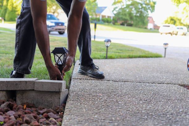 A person leans over to adjust a solar pathway light next to a concrete sidewalk and brick landscaping border.