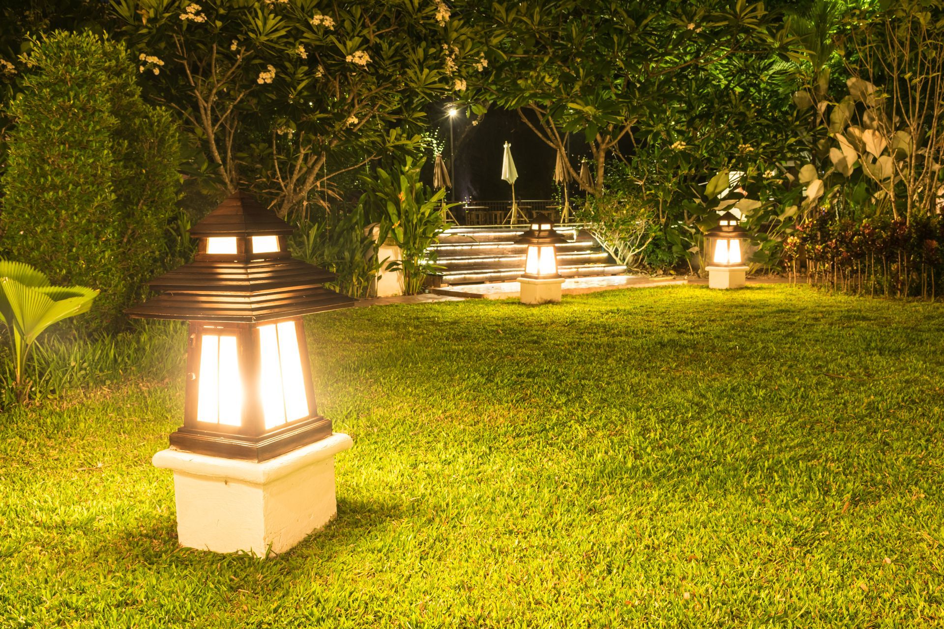 Lit garden lanterns glow in the grass, illuminating a lush backyard pathway at night.