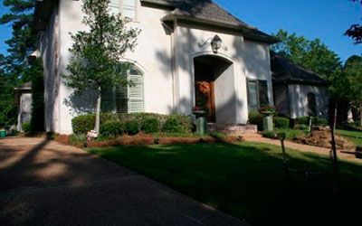 A two-story light-colored house with a dark shingled roof, arched front entryway, and a front lawn under a clear blue sky.