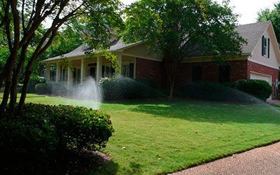 A brick house with a front porch, green lawn, and trees, with an active sprinkler watering the grass on a sunny day.
