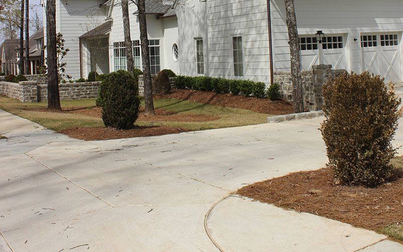 A white house with stone accents, a concrete driveway, manicured landscaping, and green trees on a sunny day.