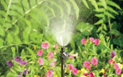 A black lawn sprinkler spraying a mist of water over a garden of pink and yellow flowers.