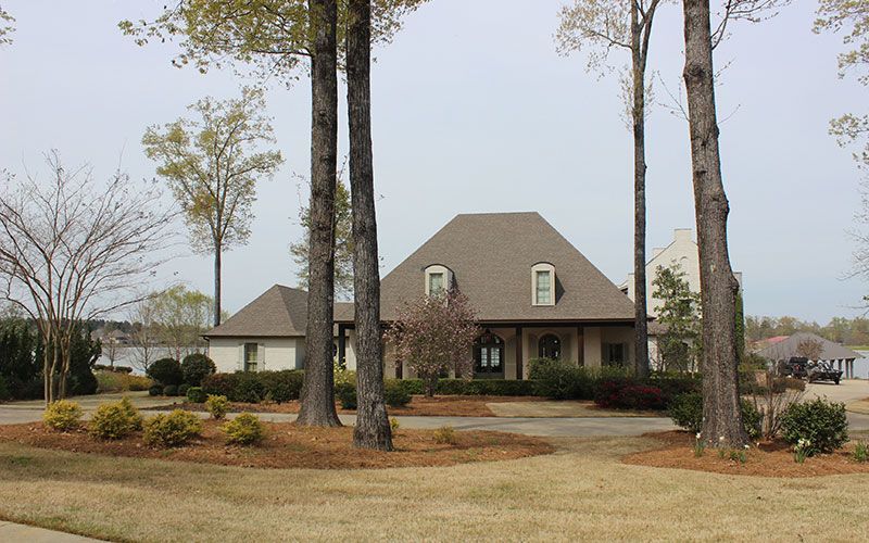 A one-story brick house with a dark roof and a covered porch, surrounded by tall pine trees and landscaping.