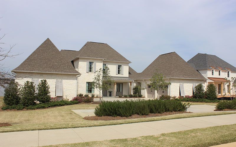 A suburban street scene showing two large, multi-story white brick houses with steep gray shingled roofs.