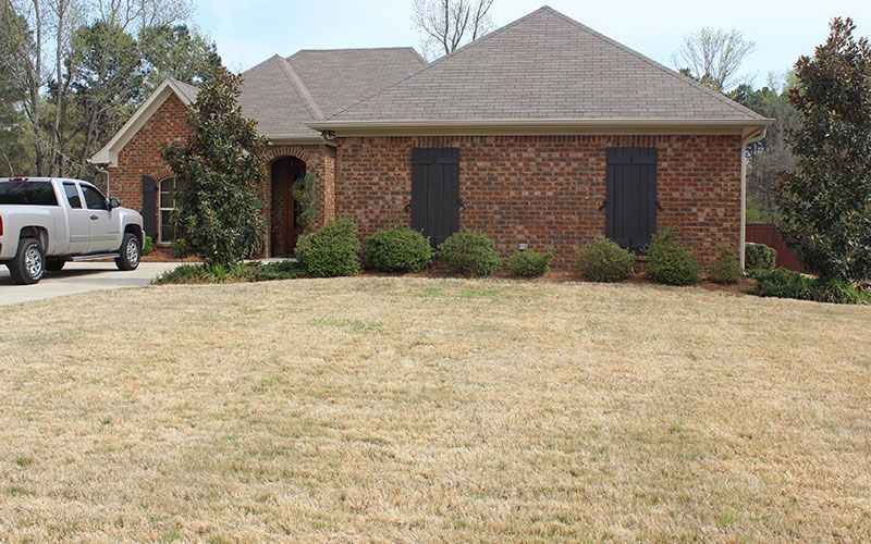 A brick house with a brown roof and dark shutters, viewed from a dry, light-brown lawn with a white pickup truck parked.