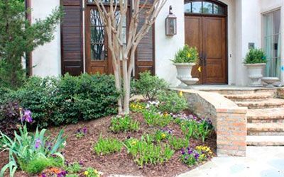 A house entrance with a stone staircase, large wooden double doors, and a front garden with a small tree and greenery.
