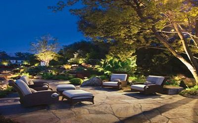 An outdoor stone patio with comfortable lounge chairs at dusk, illuminated by warm landscape lighting and trees.