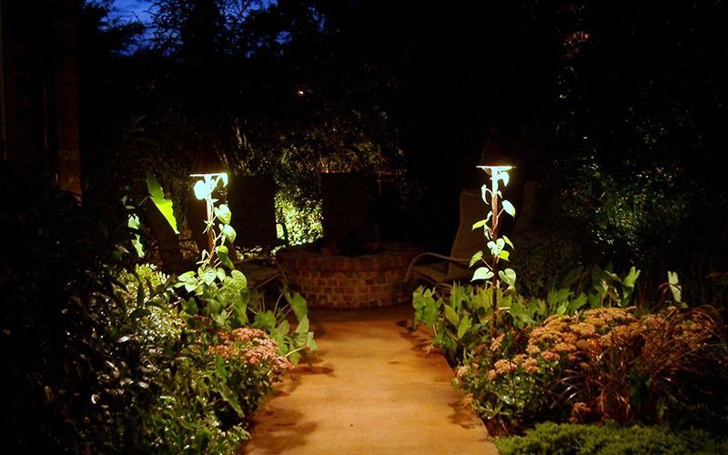 A glowing path leads through a lush garden at night, illuminated by two tall, warm-toned garden lights.