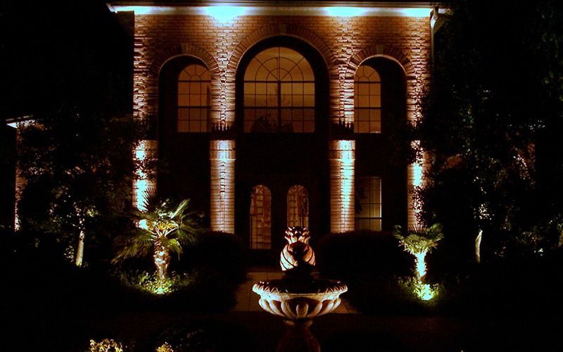A two-story brick home illuminated at night with warm landscape lighting and a central fountain in the foreground.