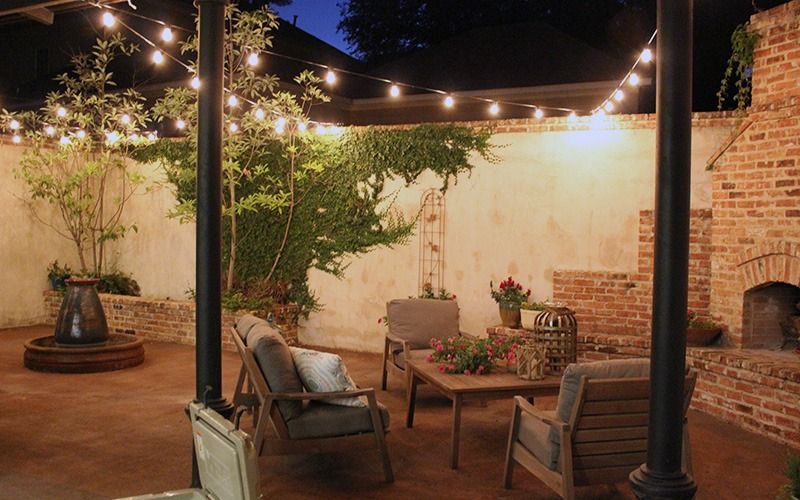 A cozy brick patio at night, illuminated by string lights, featuring two armchairs, a coffee table, and a small fountain.