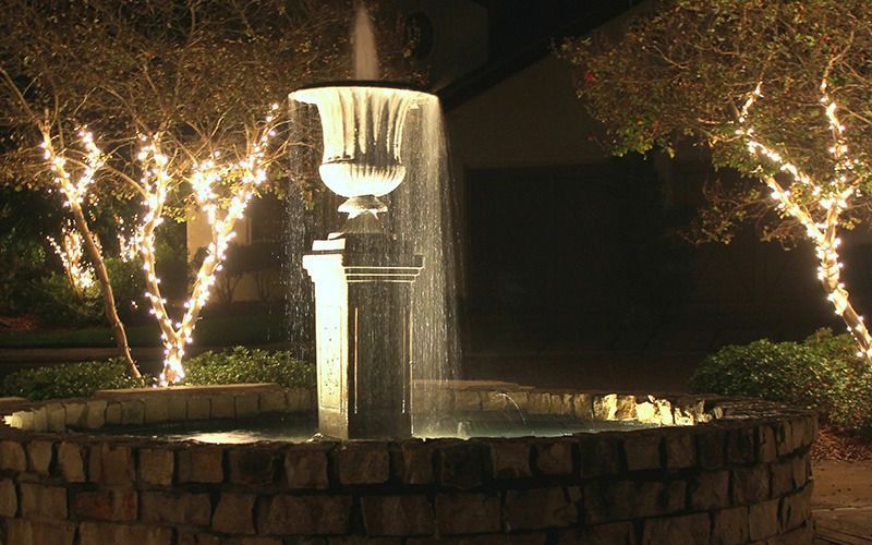 A stone fountain lit at night with water spraying into a round basin, flanked by two trees wrapped in glowing fairy lights.