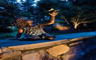 A bronze statue of a child sitting on a stone wall at night, reaching up toward a small bird perched on their hand.