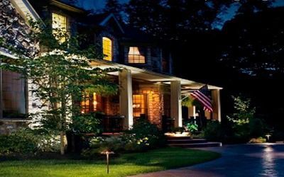 A stone house with an illuminated front porch and an American flag at dusk, surrounded by manicured landscaping.