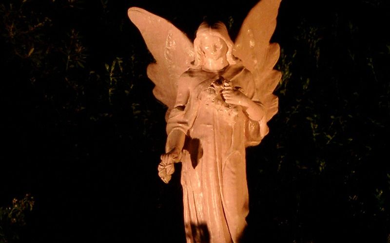 A stone angel statue illuminated against a dark background, holding a small bouquet of flowers.