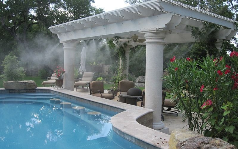 A misting system sprays water under a white pergola next to a blue swimming pool with stone accents and lounge chairs.