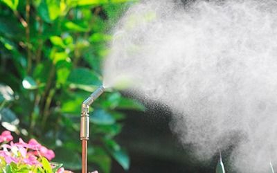 A metal nozzle sprays a fine mist of water over green plants and pink flowers in a garden.