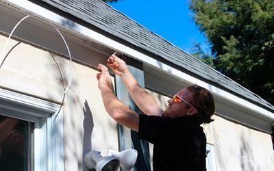 A person wearing safety glasses stands on a ladder, installing or adjusting wires near the roofline of a building.
