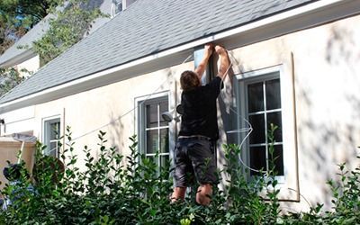 A person wearing a black shirt and shorts works on the exterior wall of a light-colored house near a window and greenery.