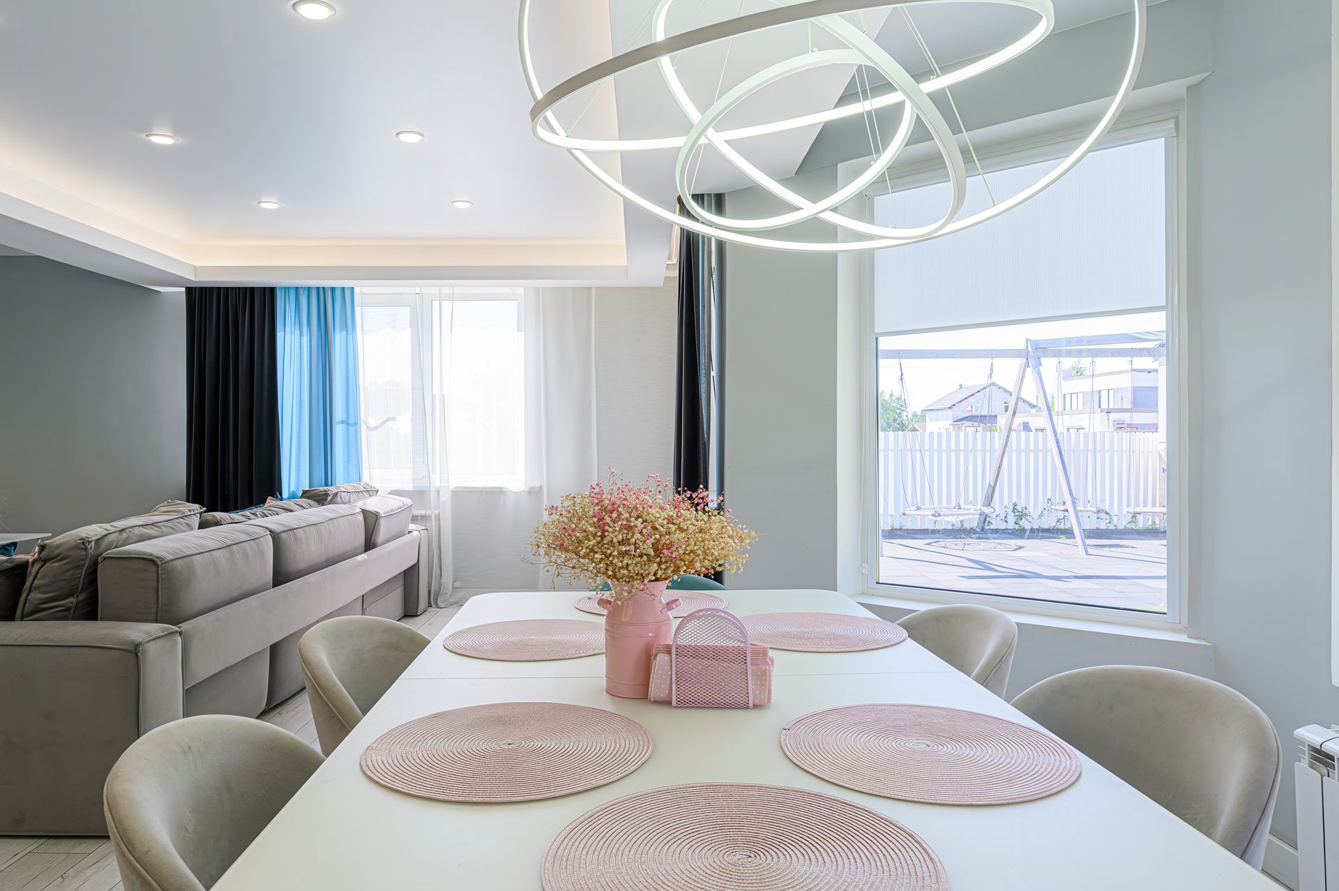 Modern dining room with white table set for six, pink floral arrangement, and view of backyard.