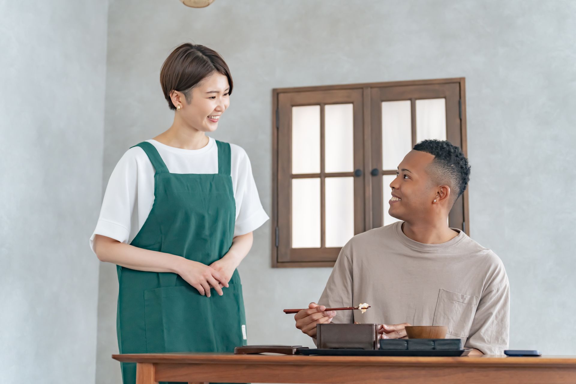 Woman in apron smiles at man eating at table. Wooden window.
