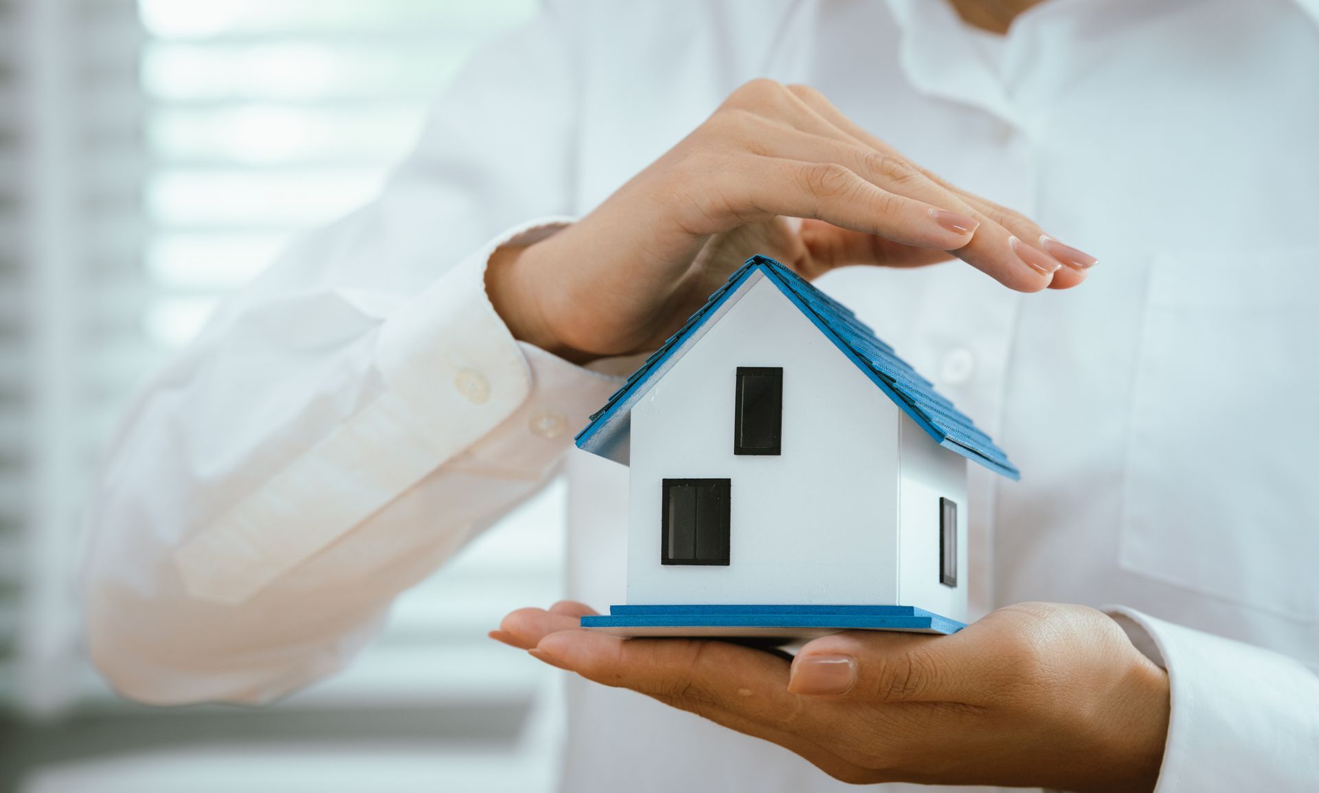 Hands cradling a miniature white house, with a blue roof, offering protection.