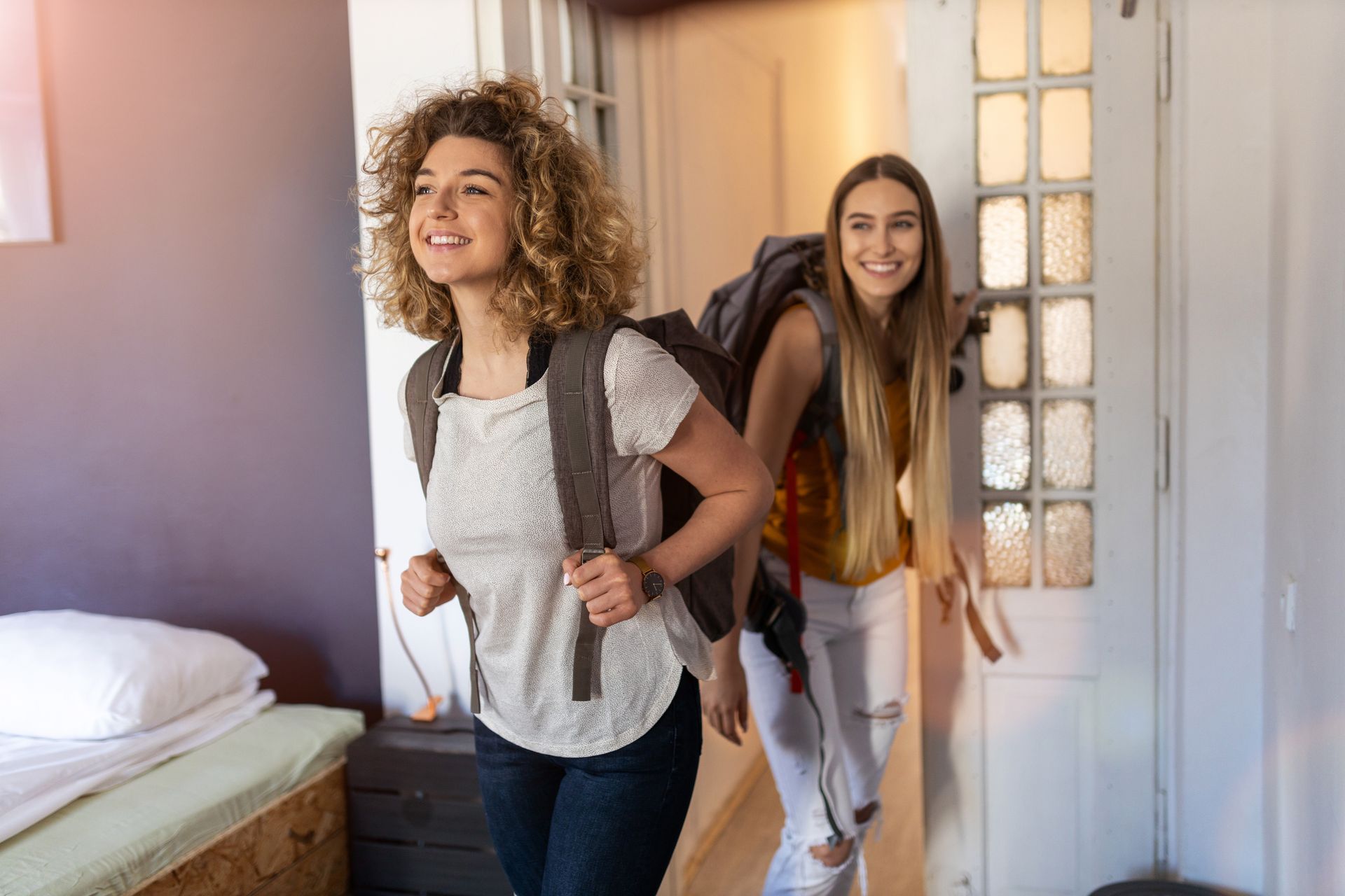 Two young women with backpacks smiling, entering a room from a doorway.
