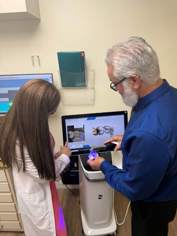 Dos personas observan una tomografía dental. Una persona con camisa azul señala una pantalla, mientras que otra con bata toca un dispositivo.