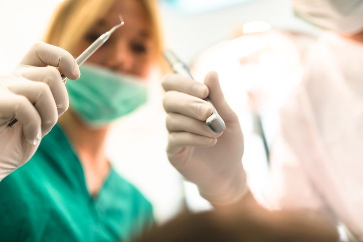 Dentist holding tools, with assistant in green scrubs; dental office setting.