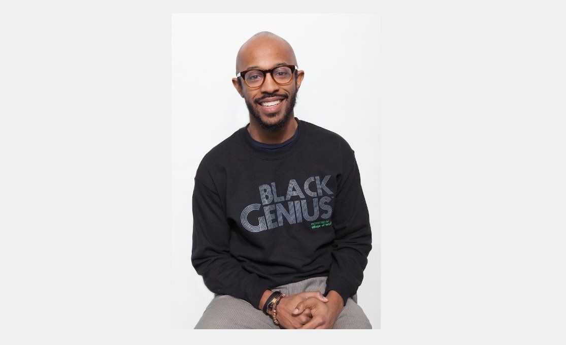 Will Jackson sits on a stool posing for a photo shoot with white background and black tshirt with slogan Black Genius protected by Village of Wisdom