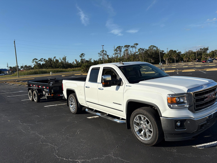 A white pickup truck parked in an empty asphalt lot while towing a black dump trailer on a sunny day.