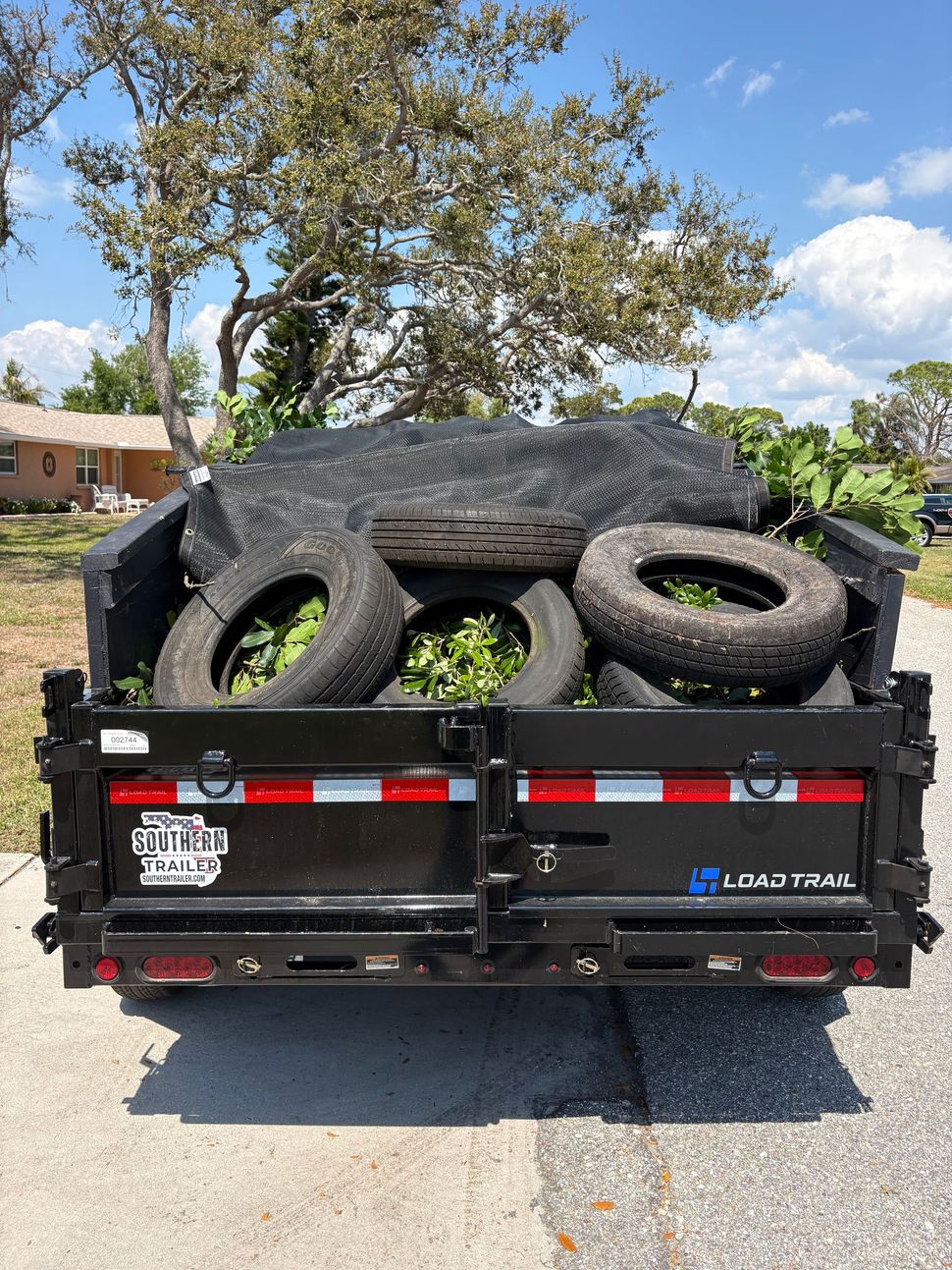 A black Load Trail dump trailer loaded with tires and yard waste parked on a residential street.