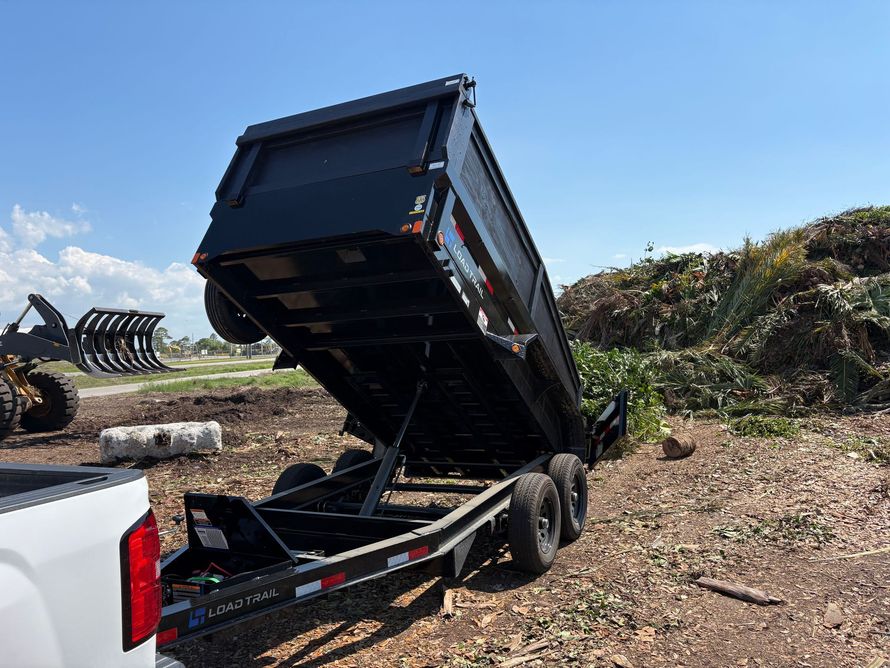 A black dump trailer raised in the dumping position at an outdoor site with a pile of yard waste in the background.