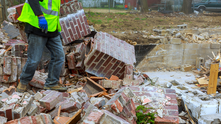 A construction worker in a high-visibility vest stands amidst a pile of red bricks and rubble at a demolition site.