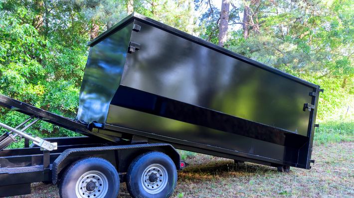 A black dumpster trailer positioned on a dirt path in a wooded area, partially tilted upward.