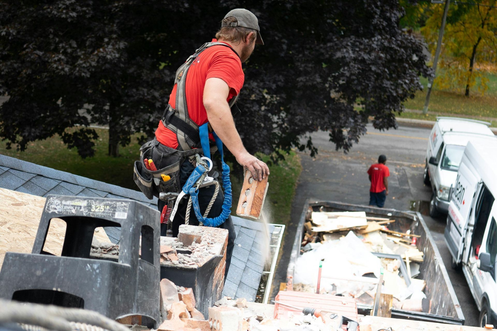 A worker in a red shirt on a roof tosses bricks into a dumpster, while a second person stands nearby on the ground.