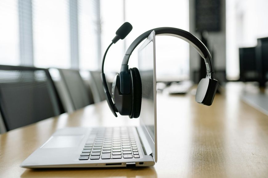 A black headset draped over the screen of an open laptop on a wooden table in a brightly lit office setting.