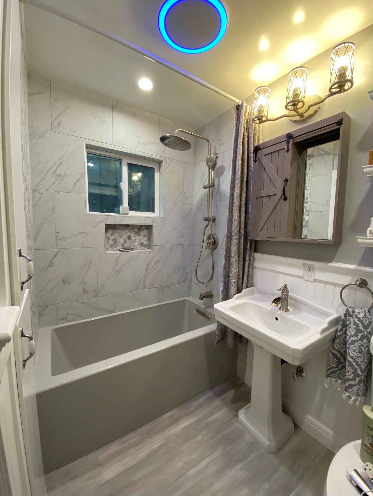 Bathroom with gray tub, marble tile, pedestal sink, and rustic medicine cabinet.