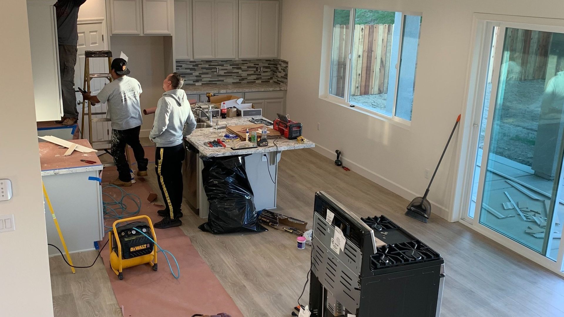 Two people renovating a kitchen, tools and materials scattered.  Light wood floors, grey cabinets, and a window/sliding door.