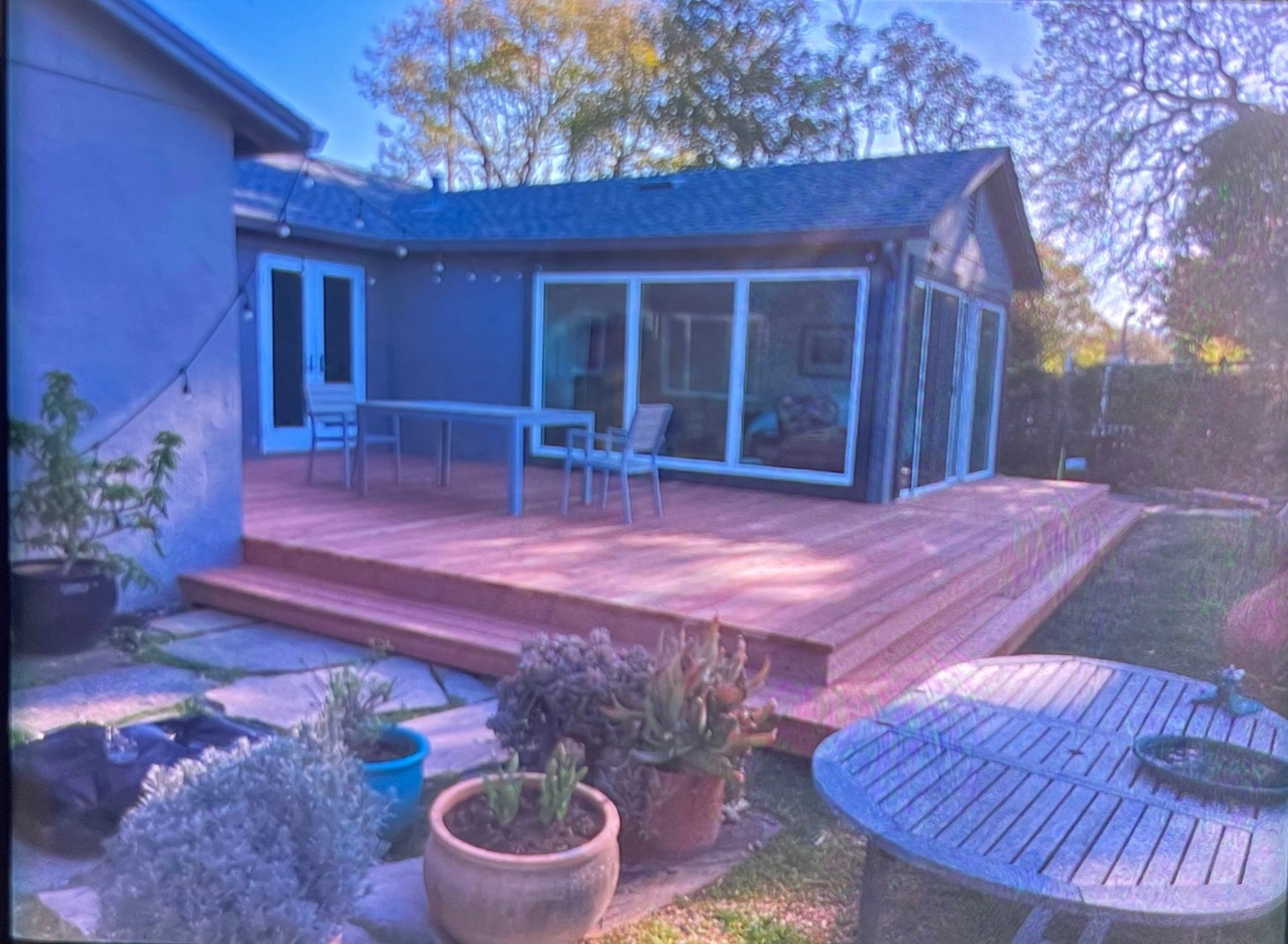 Exterior view of a blue house with a wooden deck and large windows. Potted plants in the foreground.