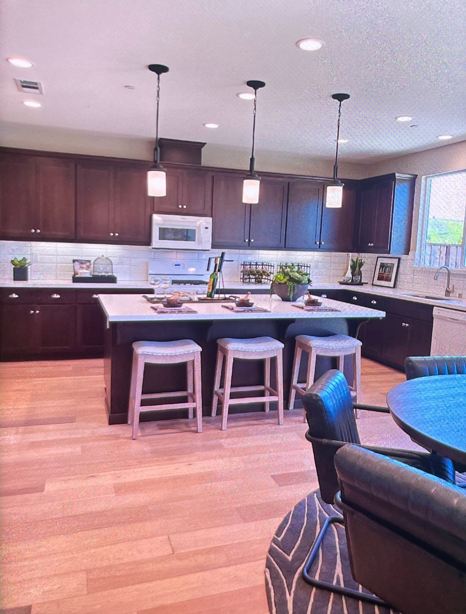 A modern kitchen with dark cabinets, white countertops, and an island with stools; pendant lights hang above.