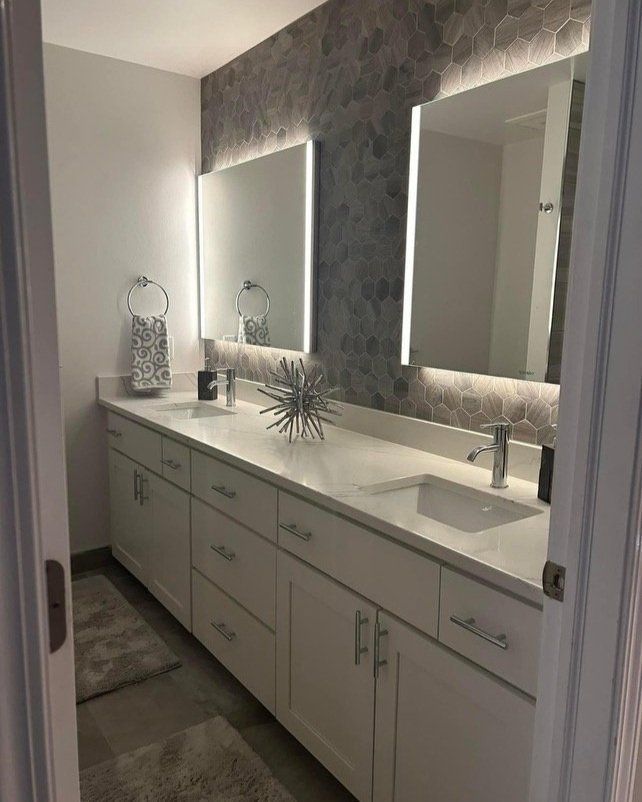 Modern bathroom with white vanity, two backlit mirrors, gray hexagon tile wall, and silver fixtures.
