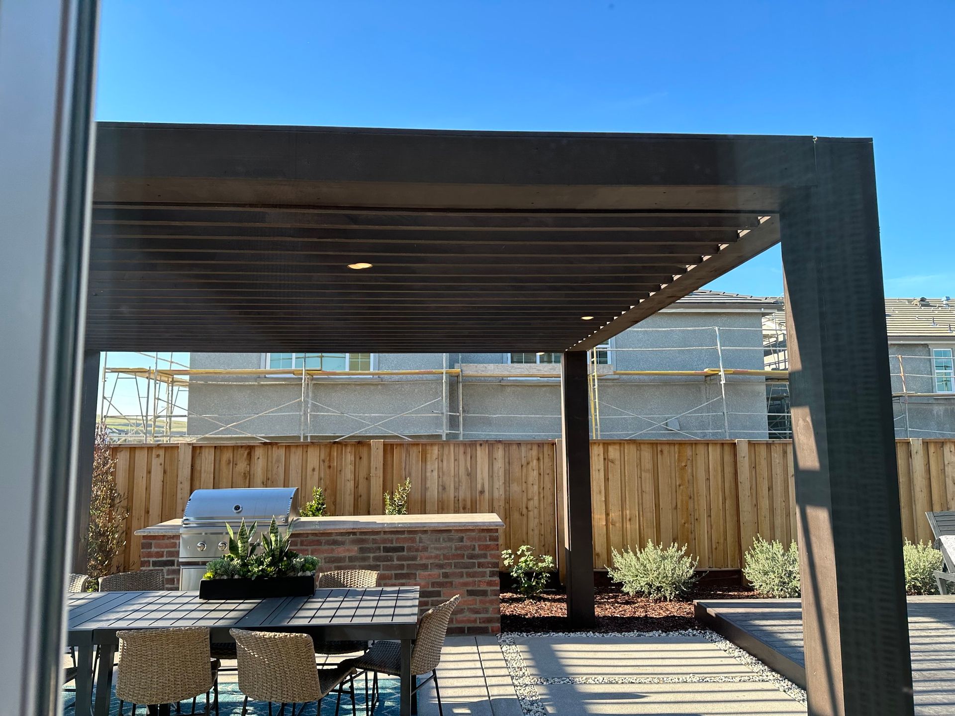 Outdoor patio with a pergola, dining set, and grill. Wooden fence and blue sky in the background.