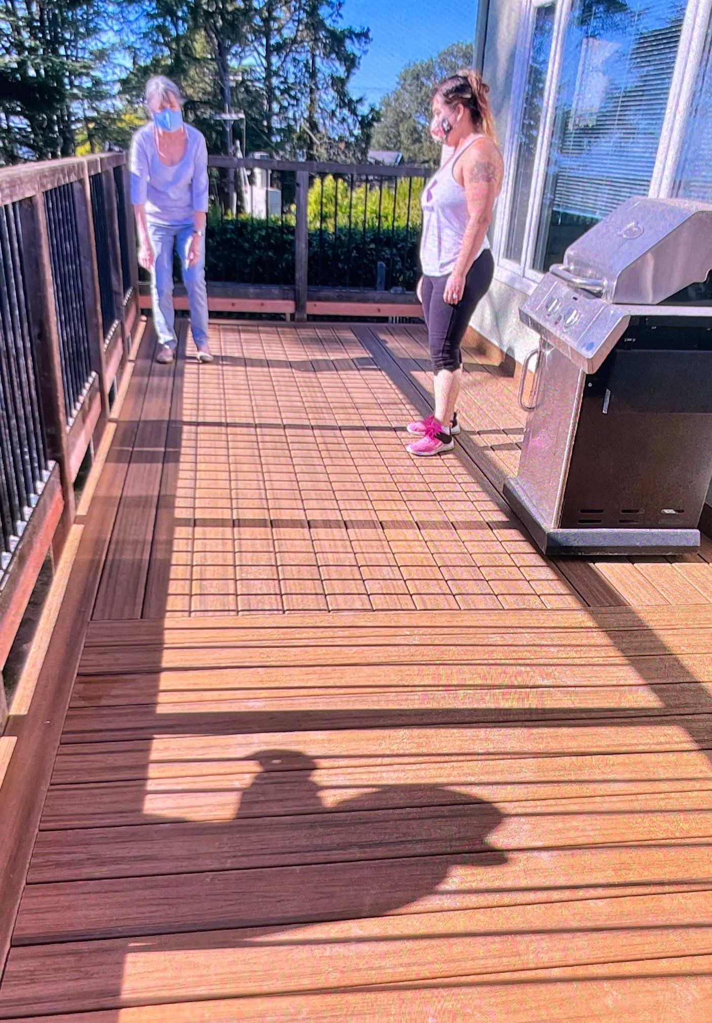 Two women on a wooden deck near a grill. One wears a mask, the other a tank top, both are smiling.
