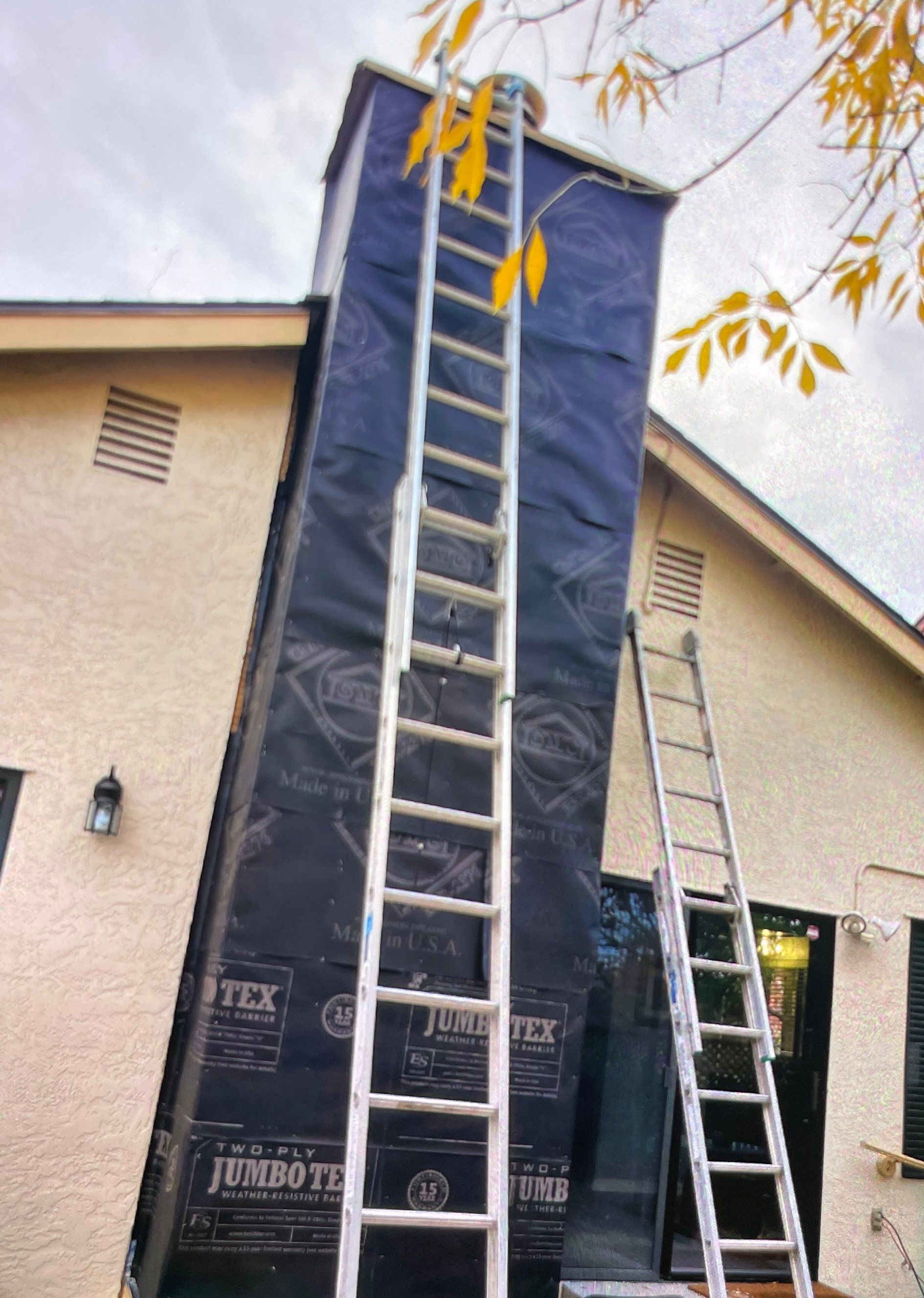 Two ladders propped against a chimney wrapped in black material; yellow leaves in the background.