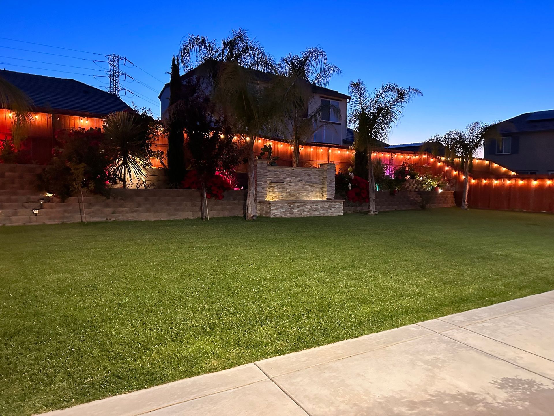 Lawn with illuminated string lights on a fence and home, with a water feature. Evening setting, blue sky.