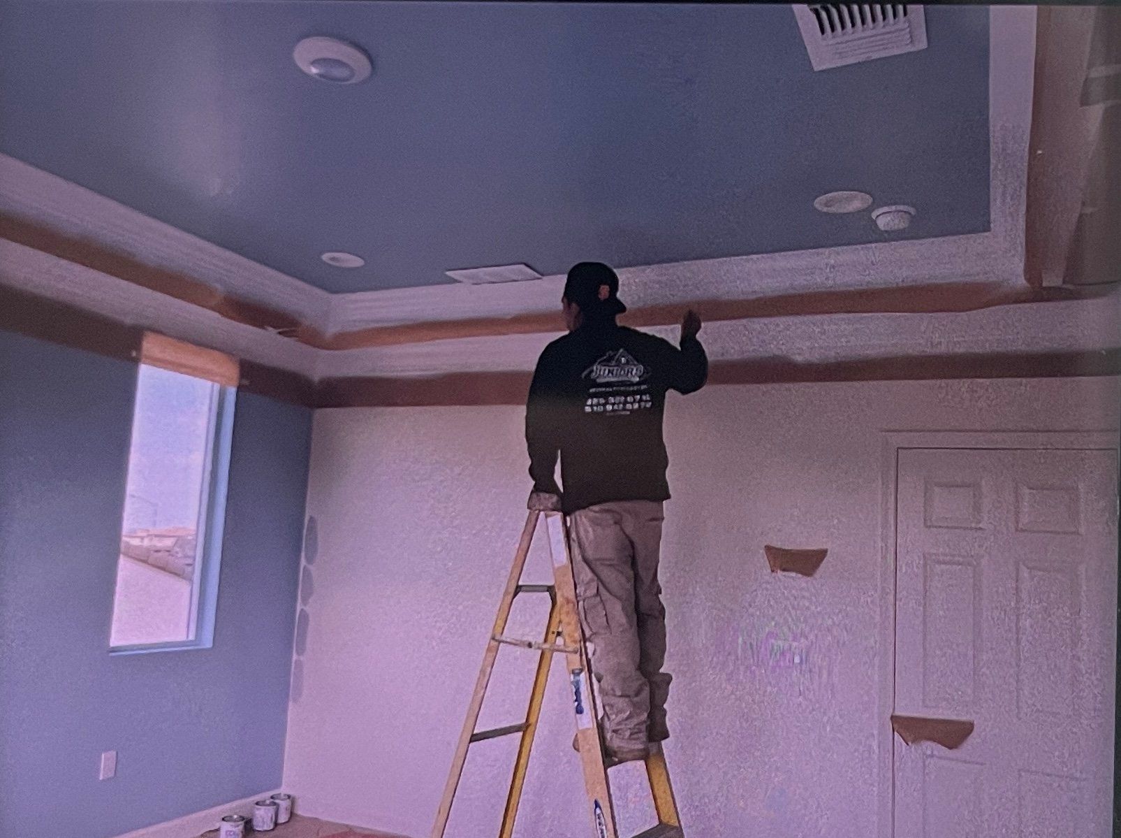 A person on a ladder paints a room's crown molding and ceiling blue. Interior painting in progress.